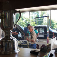 A small pile of show trophies from Greenwich, CT, to Dubai frames Wayne Carini as he regales Alfa Owners of New England with stories about finding, buying and selling cars